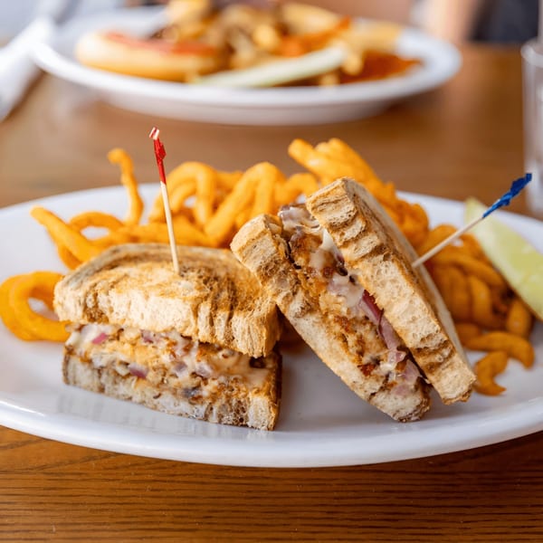 A grilled sandwich with melted cheese and meat, served with curly fries and a pickle spear.