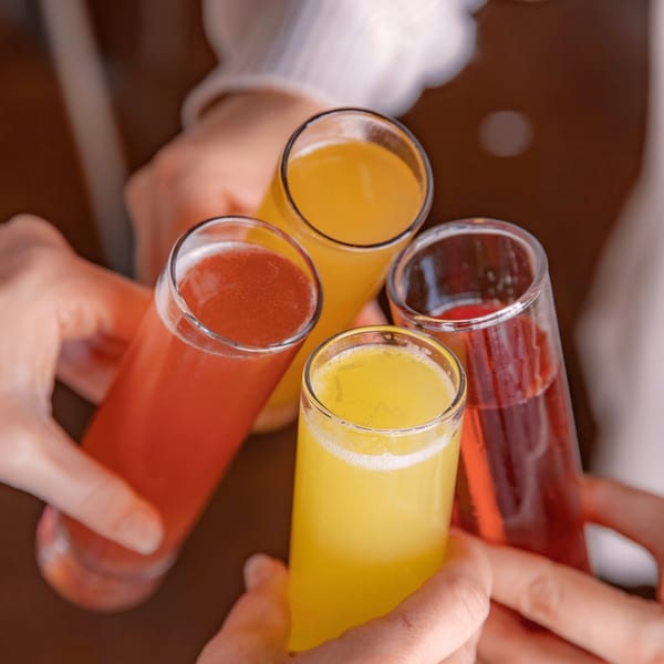 Four hands holding up glasses of colorful mimosas, two orange and two red, in a toast.