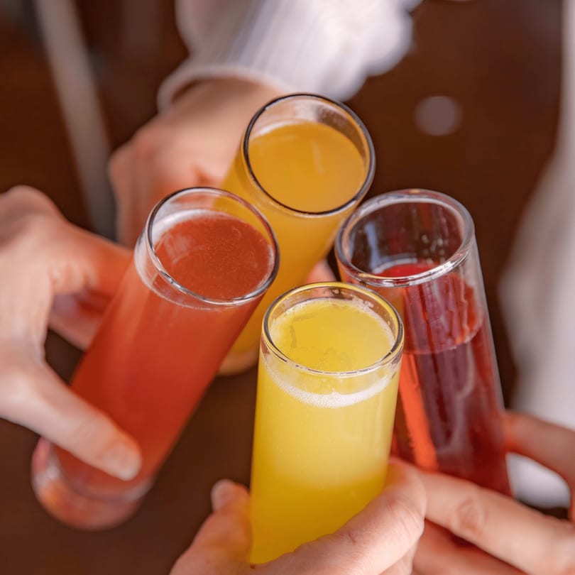 Four hands holding up glasses of colorful mimosas, two orange and two red, in a toast.