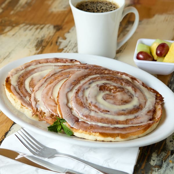 Three cinnamon roll pancakes with icing, a cup of coffee, and a side of fruit.