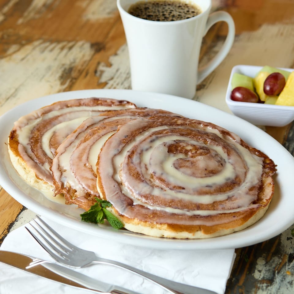 Three cinnamon roll pancakes with icing, a cup of coffee, and a side of fruit.
