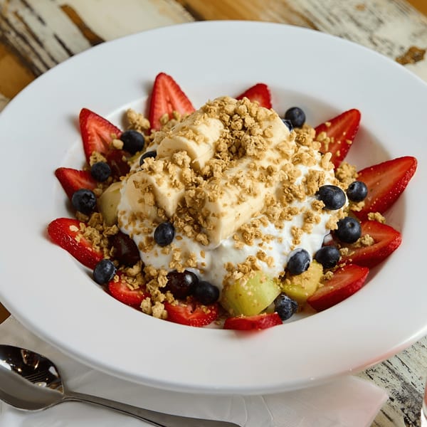 A white bowl filled with yogurt, sliced bananas, strawberries, blueberries, and granola on a wooden table.